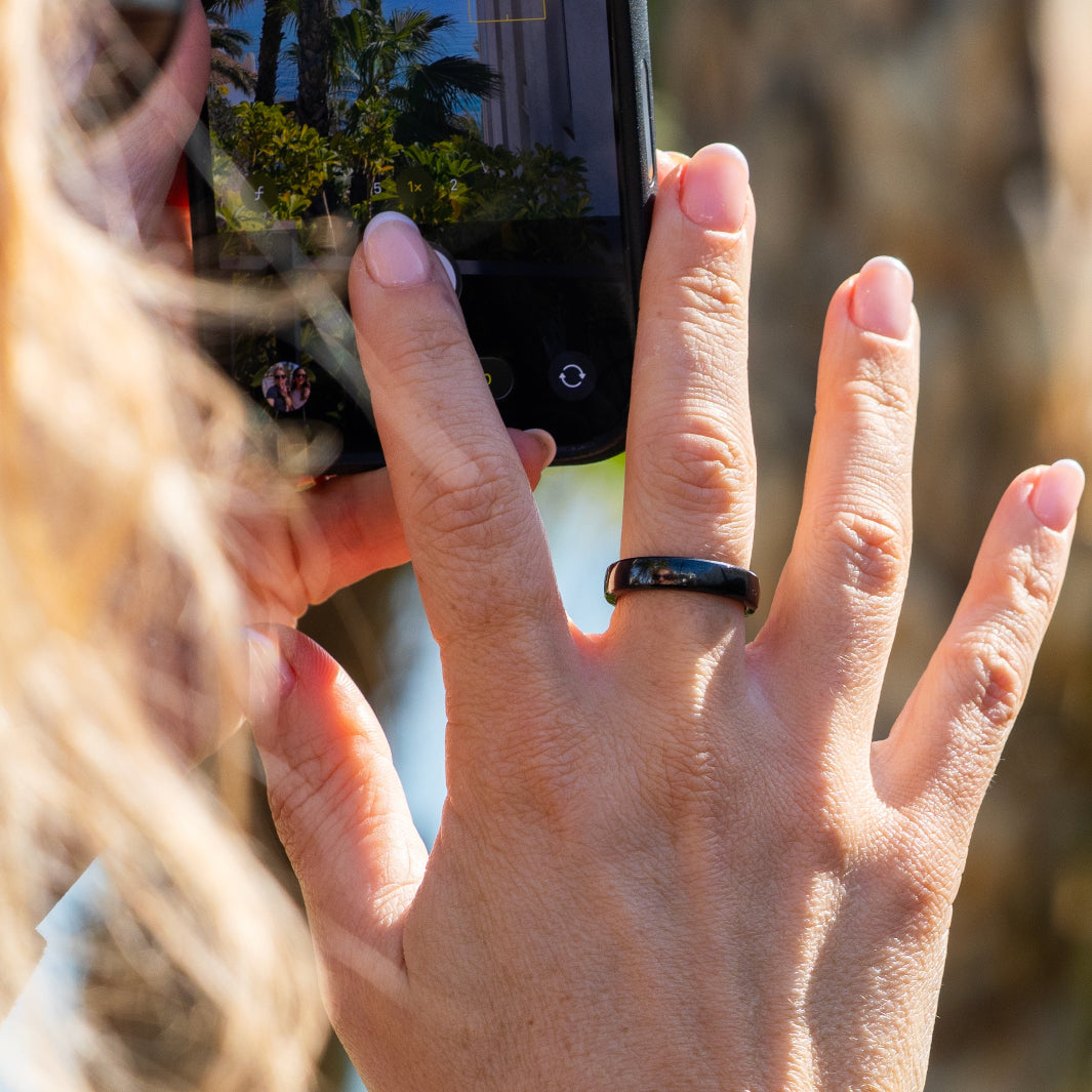 A close-up lifestyle shot of a person's hand wearing RingPay's Zirconia Ceramic Payment Ring holding a smartphone in bright outdoor sunlight.